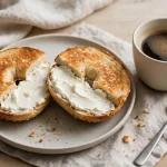 A toasted bagel spread with cream cheese on a plate next to a cup of black coffee