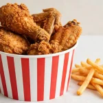 Crispy fried chicken pieces in a striped paper bucket with fries