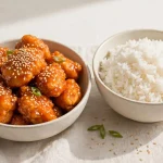 Bowl of glossy orange chicken with sesame seeds next to steamed rice
