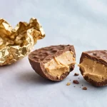A chocolate peanut butter egg candy cut in half showing the peanut butter filling, next to its gold foil wrapper
