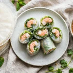 Fresh shrimp spring rolls cut in half on a plate with dry rice paper sheets and peanut dipping sauce