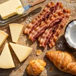 Foods high in saturated fat on a wooden board — butter, cheese wedges, bacon, coconut and croissants
