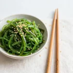 Small bowl of green seaweed salad with sesame seeds and chopsticks
