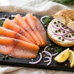 Smoked salmon slices on a board with a cream cheese bagel, capers, red onion and lemon