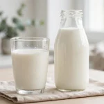 A glass of whole milk next to a glass bottle of milk on a kitchen table